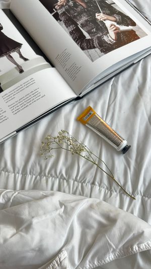 A stylish open magazine with a skincare tube and delicate flower on a white bedspread.